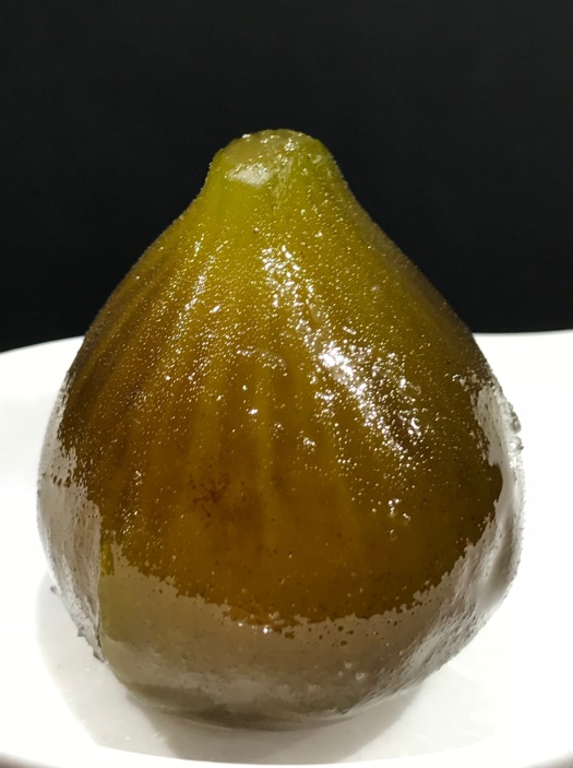 A candied unripe fig resting on a white spoon glistens with sugar syrup