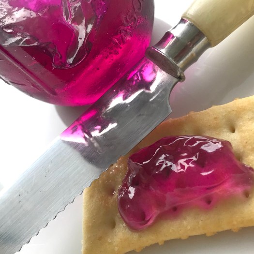 a clear glass jar showing the bright purple color of home made violet jelly, a table knife, and a golden club cracker spread with purple violet jelly